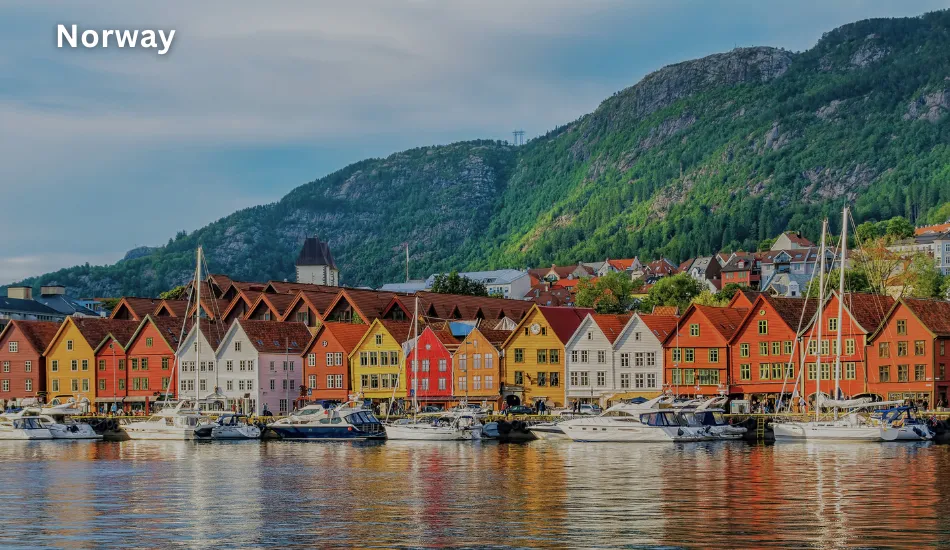 View of vibrant wooden houses and boats along the harbor in Bergen, with lush green mountains in the background, showcasing Norway as one of the best countries to migrate for free education, high quality of life, and careers in renewable energy and healthcare.